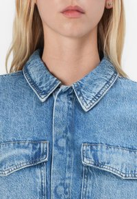 Cămașă din denim de culoare bleu deschis cu guler ascuțit, două buzunare pe piept și cusături contrastante. Materialul prezintă o textură uzată.