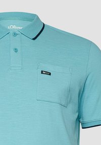 Polo bleu clair à manches courtes avec patte à deux boutons, poche poitrine avec étiquette logo et finitions foncées sur le col et les bords des manches.
