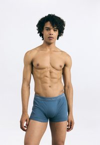 Zili kokvilnas bokseri zili ar pelēku jostasvietu, kas rotāta ar "Calvin Klein" zīmolu, uz modeļa bez kreklā.