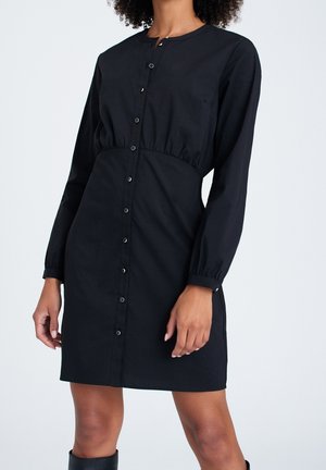 Nainen, joka pukeutunut pitkähihainen musta nappikiinnitteinen mekko, jossa on vyötäröltä kiristetty ja pyöreä pääntie, yksinkertaisella taustalla.