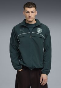 Dunkelgrüne Zip-Jacke mit weißen Akzenten, mit einem Logo auf der Brust und elastischen Bündchen. Über einem dunklen Shirt getragen und mit braunen Hosen kombiniert.