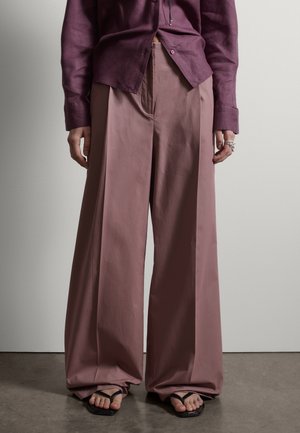 Persona con pantalones anchos color malva, camisa de botones de color púrpura oscuro parcialmente metida y sandalias negras de dedo, de pie frente a una pared gris.