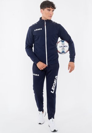 Uomo in tuta LEGEA blu scuro che tiene sotto un braccio un pallone da calcio LEGEA, in piedi davanti a uno sfondo bianco semplice.