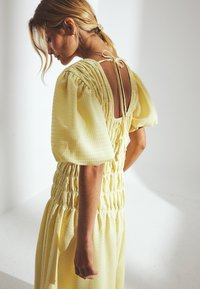 Robe jaune texturée avec des manches bouffantes, un corsage froncé et un dos ouvert avec des accents à nouer.