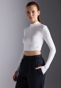 Mlada žena s crvenom kosom koja nosi bijeli dugi zip-up top s kratkim rukavima i crne hlače s vezicom, stoji s jednom rukom u džepu.