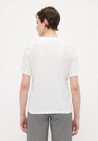 T-shirt bianco a maniche corte con scollo rotondo, texture semplice e vestibilità rilassata. Vista da dietro, abbinato a pantaloni a righe.