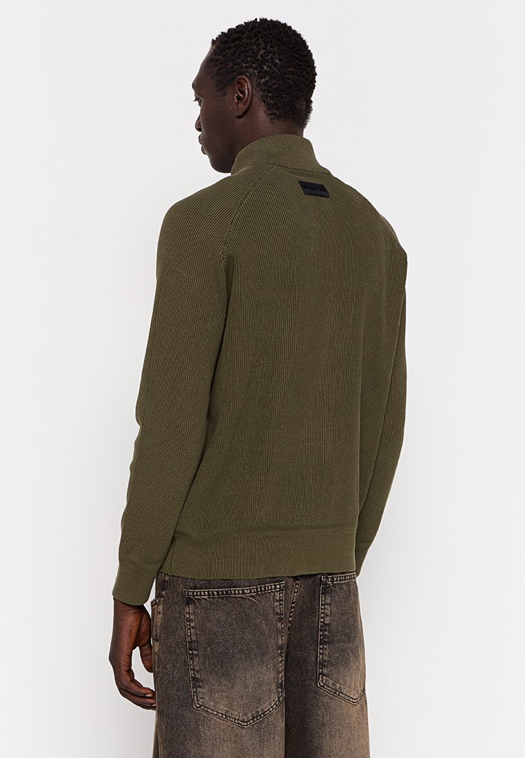 Uomo di spalle che indossa un maglione a coste di colore verde oliva e pantaloni corti in denim nero sbiadito, su uno sfondo bianco semplice.