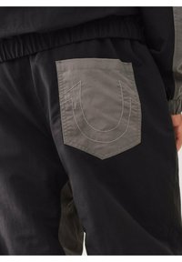 Schwarze Hose mit einer großen, grauen aufgenähten Taschen mit einem umrandeten Logo. Der Stoff wirkt glatt mit einem subtilen Glanz. Elastischer Taillenbund sichtbar.