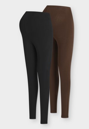 Femme enceinte en leggings marron et débardeur, debout avec un sac cabas noir sur l’épaule, portant des sandales noires devant un fond uni.