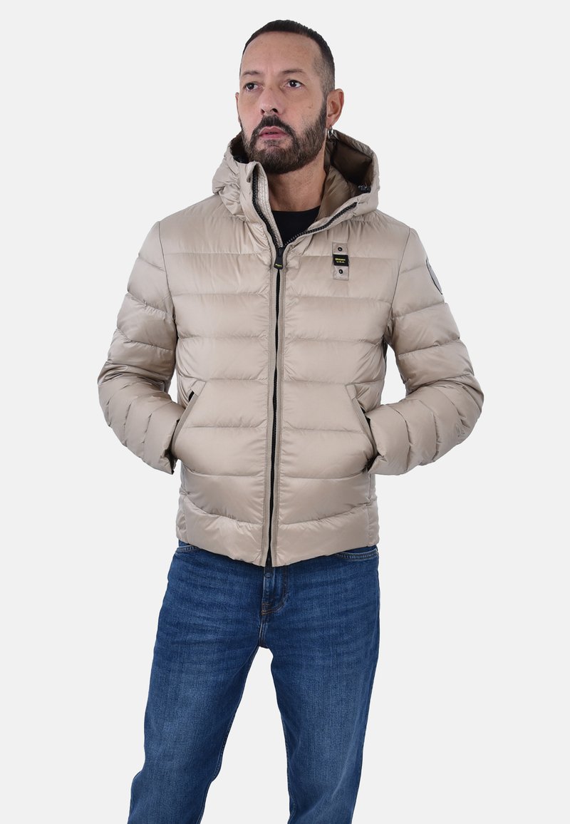 Giacca puffer beige con cappuccio, chiusura con zip e due tasche laterali, caratterizzata da un design trapuntato e un tessuto interno a contrasto.