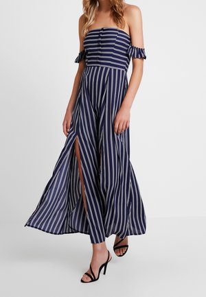 Femme portant une robe longue rayée bleu marine et blanche, épaules dénudées, avec des fentes à l'avant et des sandales noires à talons hauts.