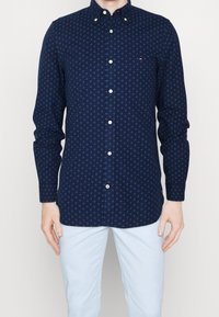 Tmavě modrá košile s knoflíky a geometrickým vzorem v modré barvě. Vyrobená z bavlny, s klasickým límečkem a řadou knoflíků. Dlouhé rukávy.