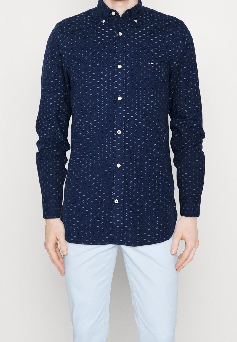 Tmavě modrá košile s knoflíky a geometrickým vzorem v modré barvě. Vyrobená z bavlny, s klasickým límečkem a řadou knoflíků. Dlouhé rukávy.