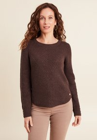 Brauner Strickpullover mit einem strukturierten Rautenmuster, gerippten Bündchen und rundem Halsausschnitt, kombiniert mit hellbraunen, figurbetonten Hosen.