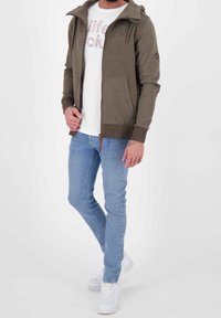 Felpa verde oliva con zip e cappuccio regolabile, indossata sopra una maglietta bianca con grafica, abbinata a jeans aderenti blu chiaro e sneakers bianche.