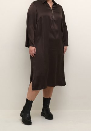 Bruine jurk met lange mouwen en een ritssluiting bij de kraag. Voorzien van zijsplitten en een gladde textuur, gecombineerd met zwarte enkellaarzen.