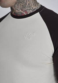 Raglan ærmet t-shirt i creme og mørkebrun, med en glat tekstur og broderet logo på brystet.