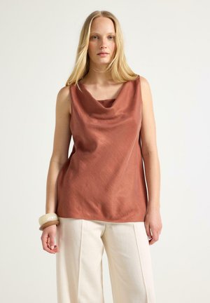 Lyshåret kvinde iført en ærmeløs top i rustfarve med draperet halsudskæring og hvide bukser med vide ben, stående mod en ensfarvet baggrund.