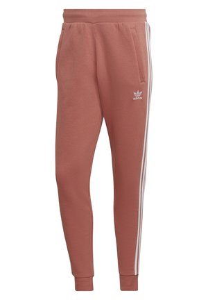 Rostfarbene Adidas Jogginghose mit elastischem Bund, weißen drei Streifen an den Seiten, Bündchen an den Knöcheln, Seitentaschen und kleinem Logo am linken Oberschenkel.