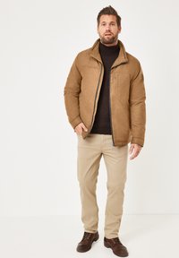 Braune strukturierte Jacke mit Reißverschluss, Stehkragen und Brusttasche; kombiniert mit beigen Hosen und braunen Lederschuhen.