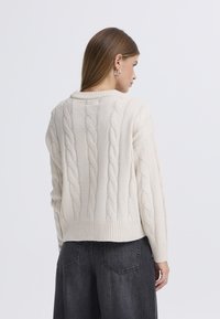 Pleteni pulover kremaste barve z vzorcem kitke, z rebrastim robom in širokimi rokavi, kombiniran s temnimi, ohlapnimi hlačami.