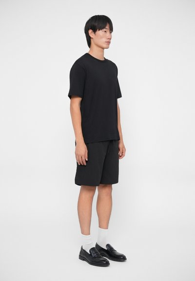 T-shirt nera in cotone, taglio ampio, abbinata a pantaloni corti neri. Scarpe nere con finitura liscia, calzini bianchi. Stile semplice e minimalista.
