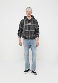 Sudadera de cuadros gris con el logo de GAP, jeans de mezclilla azul claro y zapatillas negras. El modelo se encuentra de pie contra un fondo blanco, llevando una bolsa de hombro.