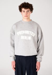 Fiatal férfi göndör hajjal, szürke kapucnis pulóverben, amelyen a "PROHIBITED BERLIN" felirat áll, sötét nadrágban, egy sima háttér előtt áll.