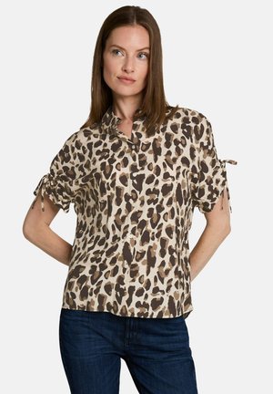 Vrouw draagt een blouse met korte mouwen en een luipaardprint, met knopen en strikjes aan de mouwen, en donkerblauwe spijkerbroek op een witte achtergrond.