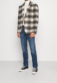 Man wearing a beige and black plaid blazer, white turtleneck, blue jeans, and black high-top sneakers standing against a white background.