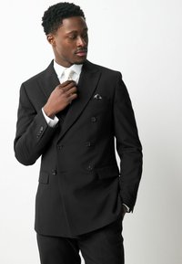 Chaqueta de traje negra de doble botonadura con botones brillantes, un pañuelo de bolsillo y una camisa blanca. Tejido suave con un corte ajustado.