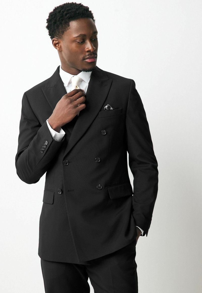 Chaqueta de traje negra de doble botonadura con botones brillantes, un pañuelo de bolsillo y una camisa blanca. Tejido suave con un corte ajustado.