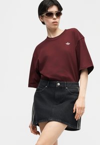 Άτομο που φοράει καφέ κοντομάνικη μπλούζα Adidas, μαύρη τζιν φούστα με πλαϊνές ρίγες και καφέ γυαλιά ηλίου, ποζάροντας μπροστά σε απλό ανοιχτό φόντο.