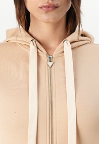 Bežna zip-up kapuca s podstavljenom kapuljačom, koja ima kremaste vezice, metalni zatvarač i glatku, meku teksturu.
