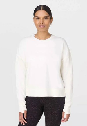 Femme aux cheveux foncés attachés en arrière, portant un haut blanc à manches longues et des leggings foncés à motifs, debout devant un fond clair uni.