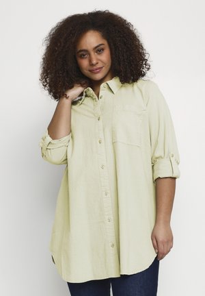 Kvinde med krøllet hår iført en lysegrøn skjorte med lange ærmer, ærmerne rullede op, smilende og rører ved sin hals.