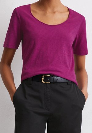 Osoba ubrana w jasnofioletowy krótki rękaw, wpuszczony w czarne spodnie z czarnym paskiem z złotą klamrą, z rękami w kieszeniach.