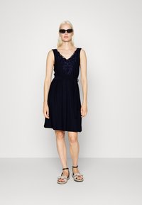 Vestido azul-marinho sem mangas com decote em V bordado, faixa na cintura e saia evasé. A modelo usa óculos de sol pretos e sandálias bege.