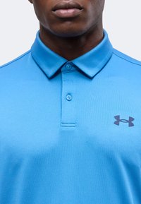 Άνδρας που φοράει μπλε πόλο Under Armour με δύο κουμπιά και γιακά, μπροστά σε απλό φόντο.