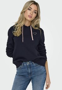 Felpa navy con lacci rosa e ricamo a forma di cuore. Realizzata in un tessuto morbido, presenta una vestibilità comoda e polsini a costine. Abbinata a jeans azzurri chiari.