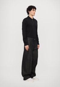 Fekete, hosszú ujjú póló, cipzáros gallérral, amelyet oversized sötét farmernadrággal párosítanak. Fehér sportcipők, minimális dizájnnal, finom textúrákkal.