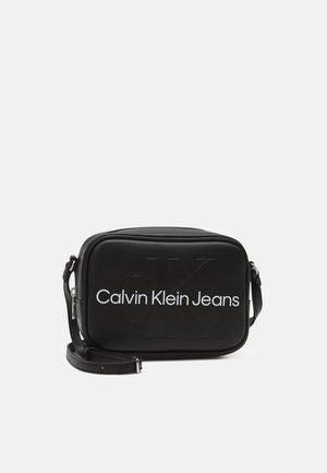 Schwarze rechteckige Umhängetasche aus glattem Material, mit geprägtem und gedrucktem "Calvin Klein Jeans"-Logo, verstellbarem Schultergurt und Reißverschluss.