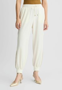 Jogging blancs avec une taille à cordon, des détails en mailles sur les côtés et des poignets élastiques. Associés à des chaussures à talons dorées pour un look contrasté.