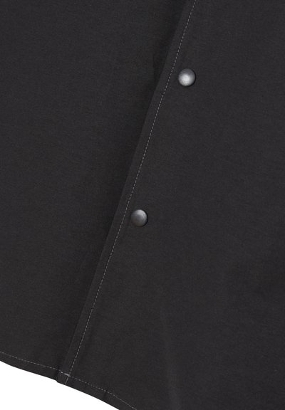 Schwarzes Kleidungsstück mit sichtbaren Nähten. Verfügt über Druckknöpfe, einen flachen Kragen und einen leicht abgerundeten Saum. Der Stoff wirkt glatt mit einem matten Finish.