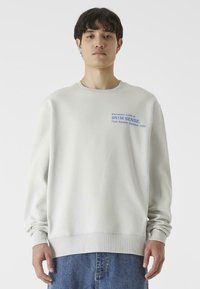 Világosszürke pulóver puha anyagból, kerek nyakkivágással, bordázott mandzsettákkal, és kék nyomtatott szöveggel a mellrészen. Kék farmernadrággal párosítva.