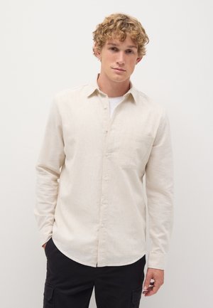 Camisa botón de color beige claro con un bolsillo en el pecho, hecha de una tela texturizada. Presenta mangas largas y un dobladillo en curva. Llevada sobre una camisa blanca.