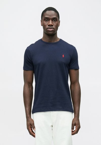 Tmavomodré bavlnené tričko s krátkymi rukávmi a okrúhlym výstrihom. Má malú červenú logo na ľavej strane hrudníka. Jemná textúra, úzky strih.