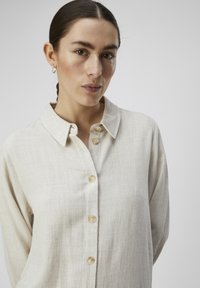 Chemise boutonée beige clair en tissu texturé, avec un col et cinq boutons sur le devant.