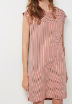 Femme portant une robe rose sans manches avec des rayures verticales blanches et noires, debout devant un fond clair uni.