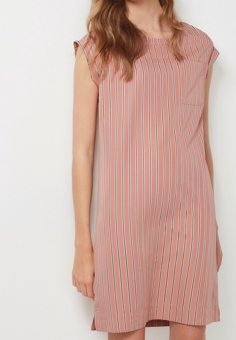Femme portant une robe rose sans manches avec des rayures verticales blanches et noires, debout devant un fond clair uni.